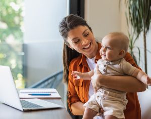 Eine Mutter sitzt vor dem Laptop und hat ein Baby im Arm.
