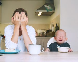 Eine Mutter stützt verzweifelt ihren Kopf in die Hände während ihr Kleinkind neben ihr sitzt und weint.