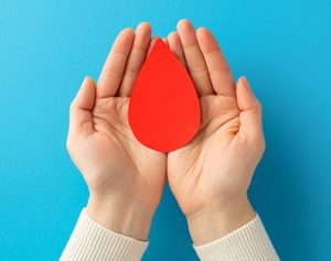 In einem aus zwei Händen geformtem Nest liegt ein Blutstropfen aus Papier.