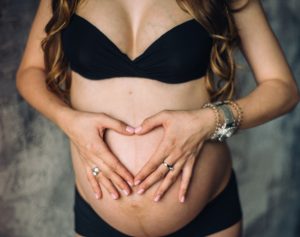 Eine Frau formt mit ihren beiden Händen ein Herz über ihrem schwangeren Bauch.