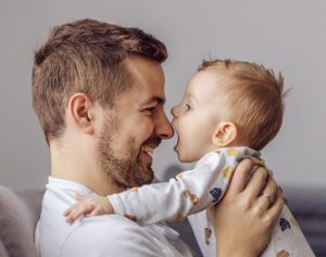 Ein Vater hält sein Baby hoch, und das Baby schnappt im Spiel nach der Nase des Vaters.