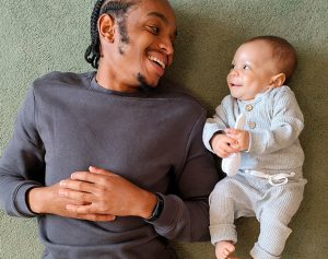 Vater liegt neben Baby und lächelt es an.