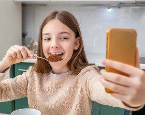 In Mädchen schiebt sich einen Löffel mit Zimt in den Mund und nimmt sich dabei mit dem Smartphone auf.