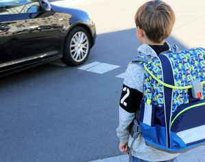 Ein Junge mit Schultasche auf dem Rücken steht am Straßenrand, während ein Auto vorbeifährt.“