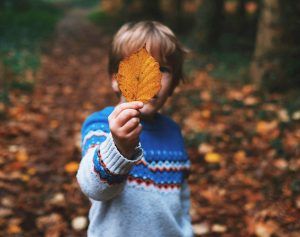 Eine Junge steht im herbstlichen Wald und hält ein Laubblatt vor sein Gesicht.