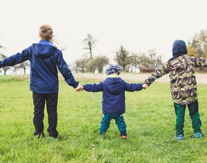 Zwei Kinder und ein Kleinkind halten sich an den Händen und stehen mit ausgestreckten Armen in einer Wiese.