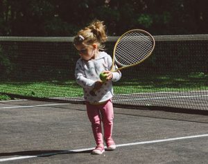 Ein Kleindkind steht mit Tennisschläger und Ball vor einem Tennisnetz.
